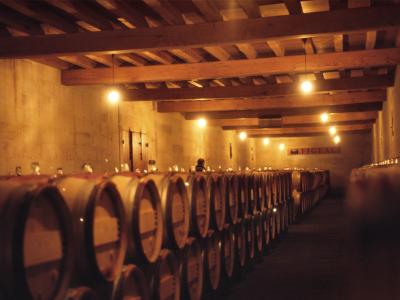 A dimly lit wine cellar with rows of wooden barrels, exposed wooden ceiling beams, and a sign on the far wall reading "FIGEAC".