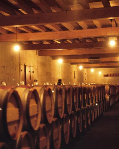 A dimly lit wine cellar with rows of wooden barrels, exposed wooden ceiling beams, and a sign on the far wall reading "FIGEAC".