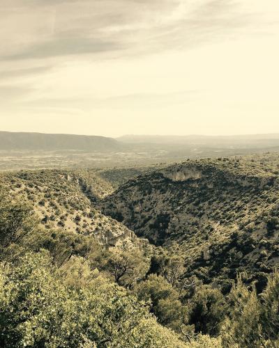 A sepia-toned landscape photograph showing a deep, scrub-covered valley or gorge with rolling hills in the foreground and a distant plateau under a hazy sky.