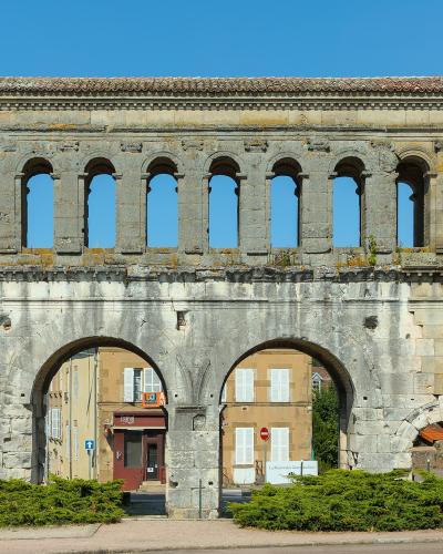 A large, ancient Roman triumphal arch with two lower arched passageways and an upper tier of ten arched openings, set against a clear blue sky with green shrubbery in the foreground and modern buildings visible through the arches.