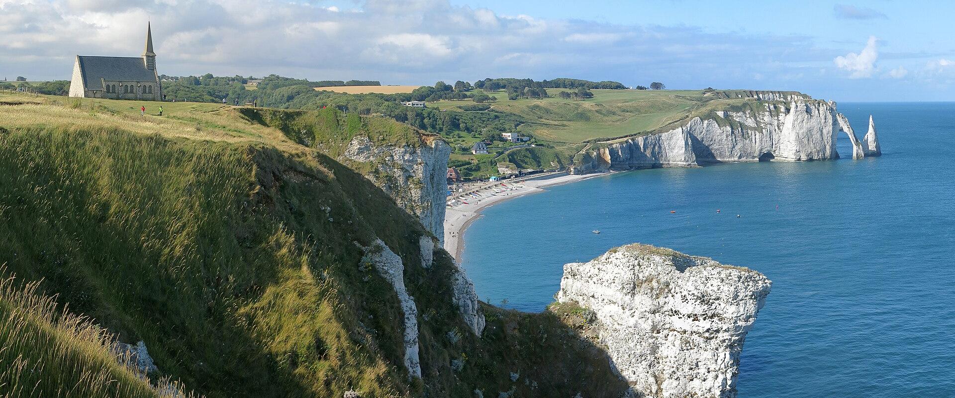 A panoramic view of the Alabaster Coast in Normandy, featuring the white chalk cliffs, the natural arch and needle rock formations of Étretat, and the Notre-Dame-de-la-Garde chapel on the cliff edge.