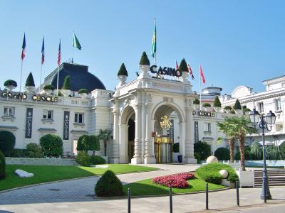 The image shows the grand entrance of the Casino Grand Cercle in Aix-les-Bains, France, identifiable by the “CASINO” and “GRAND CERCLE” signage on the building’s facade, flanked by flags and manicured gardens under a clear blue sky.