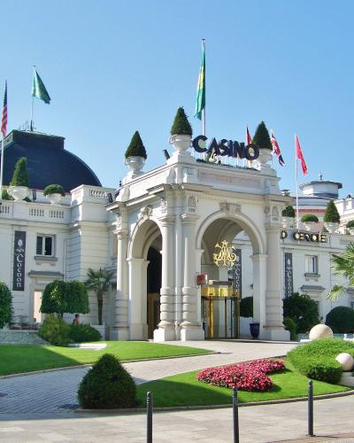 The image shows the grand entrance of the Casino Grand Cercle in Aix-les-Bains, France, identifiable by the “CASINO” and “GRAND CERCLE” signage on the building’s facade, flanked by flags and manicured gardens under a clear blue sky.