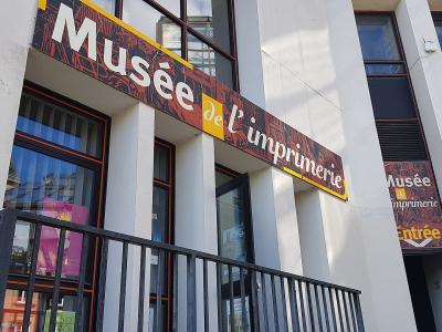 The image shows the exterior facade of a building with a large sign reading "Musée de l'imprimerie" and a smaller sign indicating "Entrée" (Entrance).