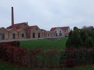 The image shows a large, historic industrial complex with stone buildings, red-tiled roofs, and a tall brick chimney, set in a grassy landscape with autumnal foliage in the foreground.