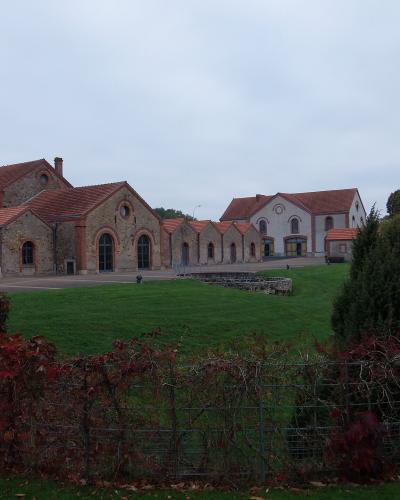 The image shows a large, historic industrial complex with stone buildings, red-tiled roofs, and a tall brick chimney, set in a grassy landscape with autumnal foliage in the foreground.