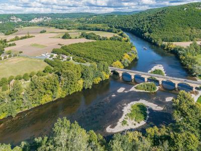 An aerial view of a multi-arched stone bridge spanning a wide river with visible sandbars, surrounded by green forests and agricultural fields under a cloudy sky.