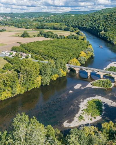 An aerial view of a multi-arched stone bridge spanning a wide river with visible sandbars, surrounded by green forests and agricultural fields under a cloudy sky.