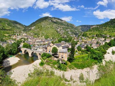 A panoramic view of the village of Sainte-Enimie nestled in a river valley, featuring a prominent multi-arch stone bridge over the Tarn River, surrounded by steep, green, forested hills under a blue sky with scattered clouds.