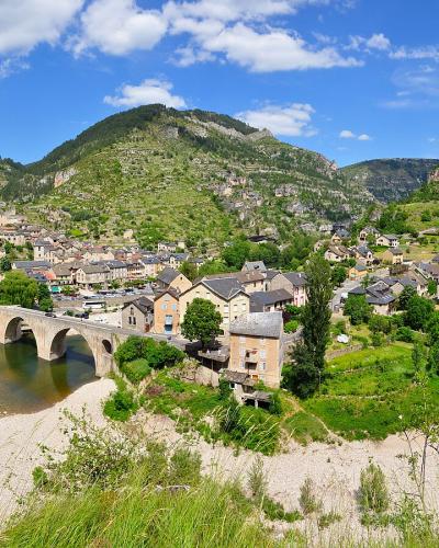 A panoramic view of the village of Sainte-Enimie nestled in a river valley, featuring a prominent multi-arch stone bridge over the Tarn River, surrounded by steep, green, forested hills under a blue sky with scattered clouds.