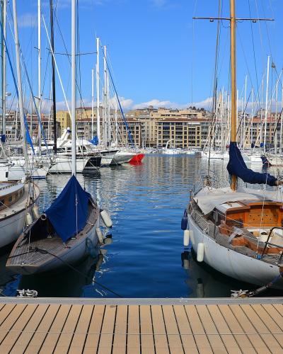 The image shows a marina filled with sailboats docked at wooden piers, with a backdrop of multi-story urban buildings under a blue sky.