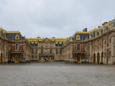 A wide-angle photograph of the Cour d'Honneur (Cour de Marbre) of the Palace of Versailles, showing the central pavilion flanked by the wings of the King's and Queen's apartments, with distinctive red brick and stone facades, blue slate roofs with go