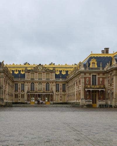A wide-angle photograph of the Cour d'Honneur (Cour de Marbre) of the Palace of Versailles, showing the central pavilion flanked by the wings of the King's and Queen's apartments, with distinctive red brick and stone facades, blue slate roofs with go