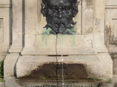 A close-up of a stone fountain featuring a bronze mask spout with water flowing into a basin, set against a stone wall with a sign reading "EAU NON POTABLE".