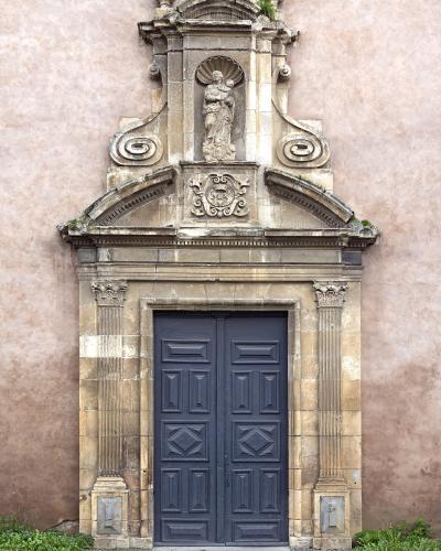The image shows an ornate, classical stone doorway set into a pinkish stucco wall, featuring fluted pilasters, a pediment with a cartouche, and a niche above containing a statue of the Virgin and Child.