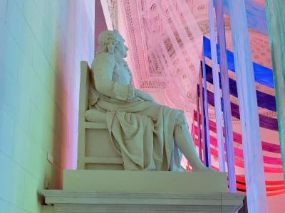 A white marble statue of a seated man in 18th-century attire, positioned on a pedestal inside a building with an ornate coffered ceiling and modern colorful fabric installations.