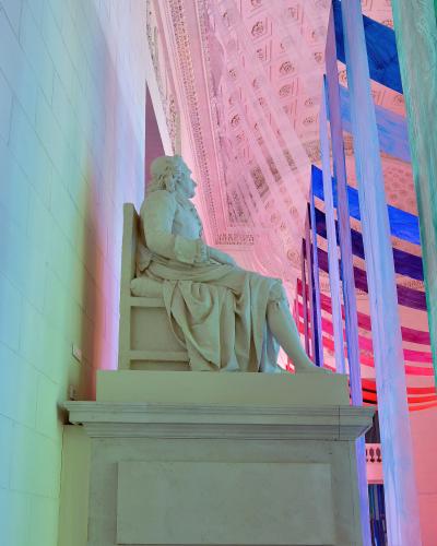 A white marble statue of a seated man in 18th-century attire, positioned on a pedestal inside a building with an ornate coffered ceiling and modern colorful fabric installations.