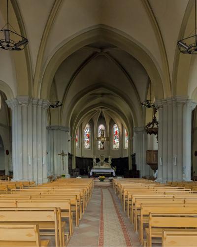 The interior nave of a church with light wood pews, white columns, vaulted ceilings, and stained glass windows at the altar.