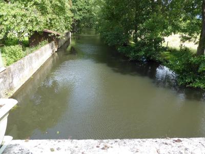 The image shows a calm, narrow river or canal flanked by a weathered stone retaining wall on the left and dense green trees on the right, with a stone parapet visible in the immediate foreground.