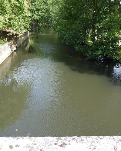 The image shows a calm, narrow river or canal flanked by a weathered stone retaining wall on the left and dense green trees on the right, with a stone parapet visible in the immediate foreground.