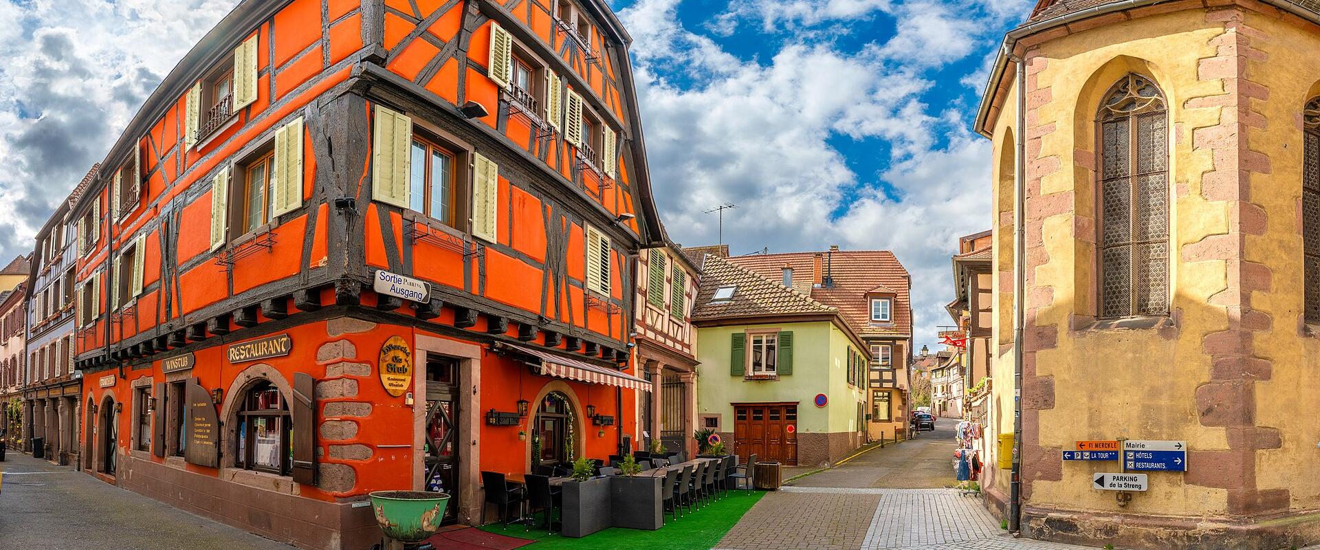 1. A vibrant orange half-timbered building labeled “Restaurant” and “Sofia’s Bistro” stands at a cobblestone street corner in a historic European town, with a yellow stone chapel or church tower to the right under a blue sky with scattered clouds.