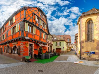 1. A vibrant orange half-timbered building labeled “Restaurant” and “Sofia’s Bistro” stands at a cobblestone street corner in a historic European town, with a yellow stone chapel or church tower to the right under a blue sky with scattered clouds.