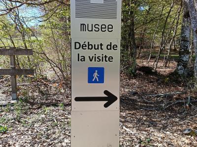A tall, vertical white signpost in a wooded area displaying a stylized hut icon, the word "musee", the phrase "Début de la visite" (Start of the visit), a blue pedestrian symbol, and a large black arrow pointing right.