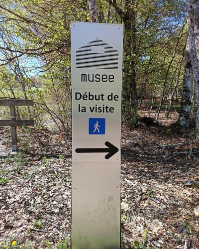 A tall, vertical white signpost in a wooded area displaying a stylized hut icon, the word "musee", the phrase "Début de la visite" (Start of the visit), a blue pedestrian symbol, and a large black arrow pointing right.
