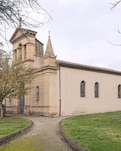 A beige stone church with a bell tower and spire, set in a grassy area with trees and a gravel path leading to the entrance.