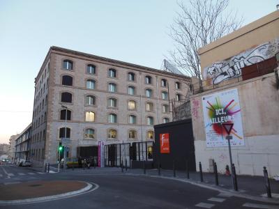 A street-level view of a large, multi-story brick industrial building with arched windows, featuring an orange sign reading "FRICHE entrée" at the gate, alongside a wall with graffiti and a poster saying "ICI, AILLEURS".