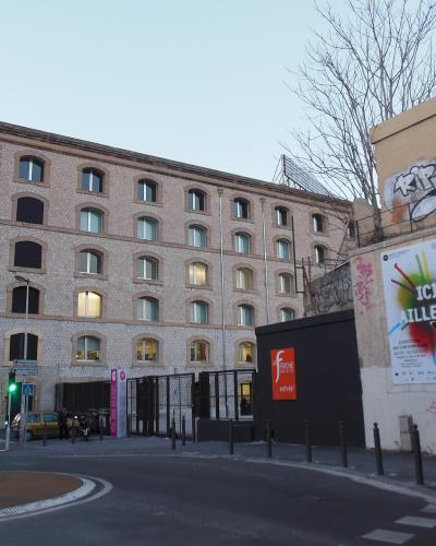 A street-level view of a large, multi-story brick industrial building with arched windows, featuring an orange sign reading "FRICHE entrée" at the gate, alongside a wall with graffiti and a poster saying "ICI, AILLEURS".