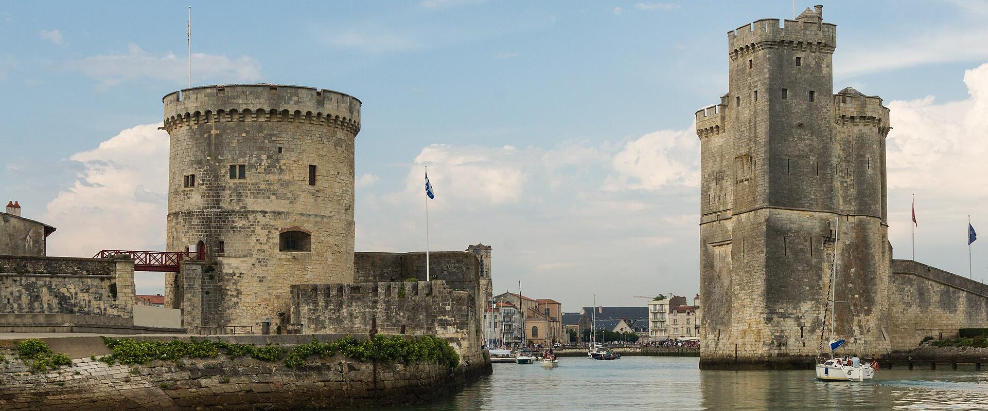 1. The image shows two historic stone towers flanking a narrow waterway entrance, with boats visible in the channel and flags flying atop the structures.