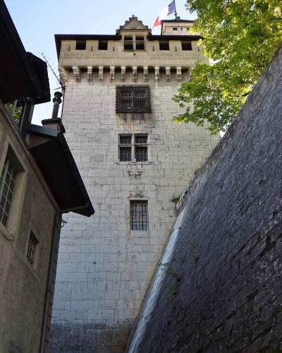 A low-angle view of a tall, light-colored stone tower with crenellations, barred windows, and a French flag flying from the roof, situated between adjacent buildings and a sloping stone wall.