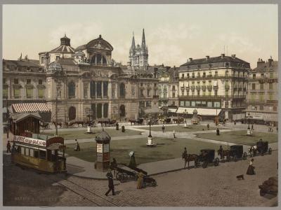 A vintage colorized photograph of a large European city square featuring a grand ornate building (likely a theater or opera house) on the left, horse-drawn carriages and an early tram in the foreground, with a cathedral spire visible in the backgr