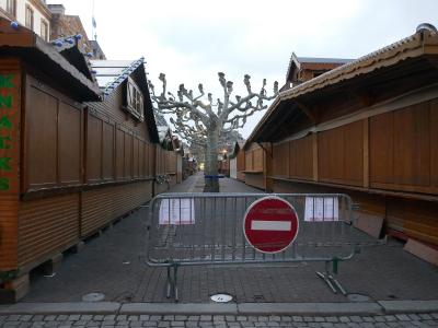 A row of closed wooden market chalets with a "KNAKKS" sign, a bare tree, and a metal barrier with a red "no entry" sign blocking access to the walkway.