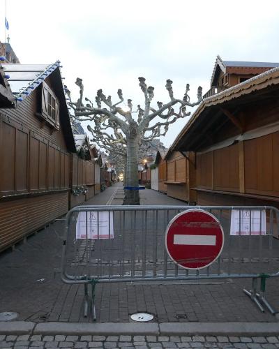 A row of closed wooden market chalets with a "KNAKKS" sign, a bare tree, and a metal barrier with a red "no entry" sign blocking access to the walkway.