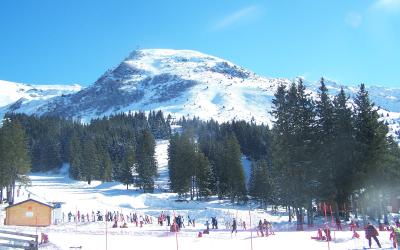 A snowy ski slope with skiers and snowboarders, evergreen trees, a small wooden building on the left, and a prominent snow-covered mountain peak in the background under a clear blue sky.