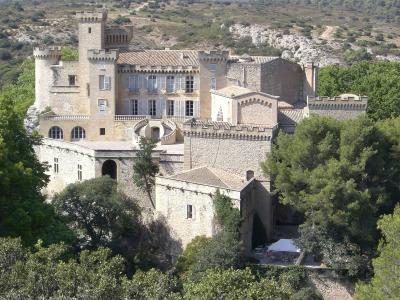 A large, fortified stone castle with crenellated towers, arched windows, and a prominent double spiral staircase, set on a hillside surrounded by green trees and dry scrubland.