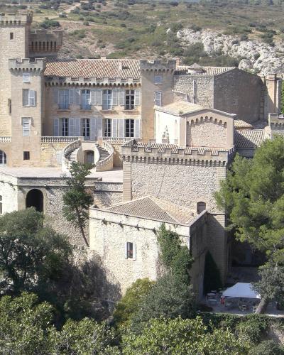 A large, fortified stone castle with crenellated towers, arched windows, and a prominent double spiral staircase, set on a hillside surrounded by green trees and dry scrubland.
