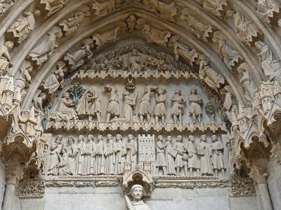 A close-up photograph of an ornate Gothic stone portal tympanum and archivolts, featuring detailed sculptural reliefs of biblical scenes and figures, with a statue of a saint in a niche below.