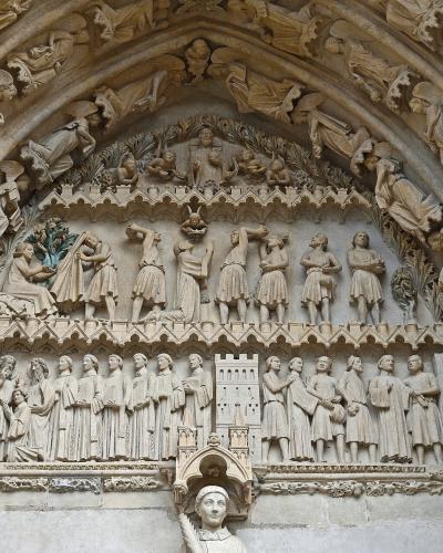 A close-up photograph of an ornate Gothic stone portal tympanum and archivolts, featuring detailed sculptural reliefs of biblical scenes and figures, with a statue of a saint in a niche below.