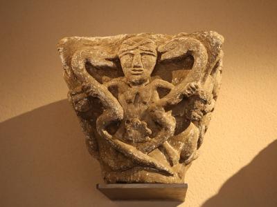 A close-up photograph of a weathered stone architectural capital featuring a carved figure entwined with serpents, displayed on a small metal stand against a plain wall.