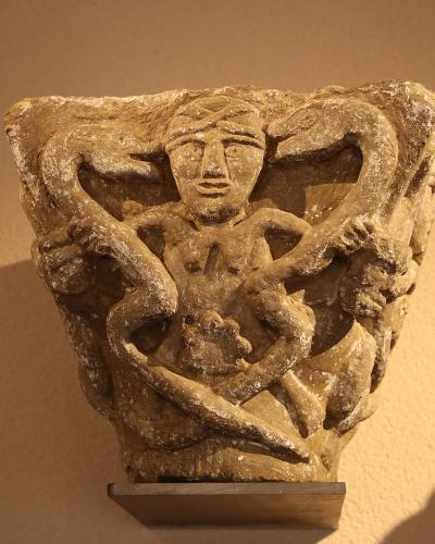 A close-up photograph of a weathered stone architectural capital featuring a carved figure entwined with serpents, displayed on a small metal stand against a plain wall.