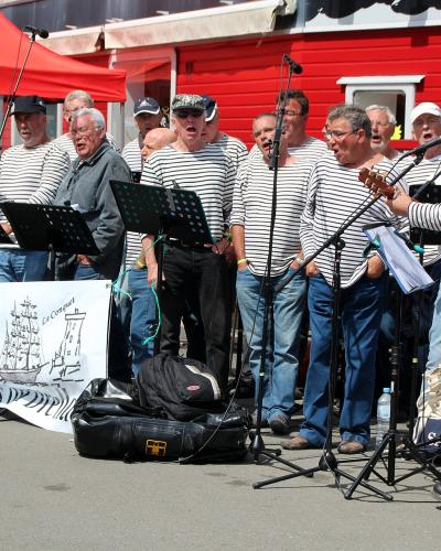 A group of men in striped sailor shirts performing outdoors on a paved area, with a banner displaying "Le Conquet" and a ship illustration, alongside a "Groupama" sponsor sign and a red building labeled "Bistro" in the background.