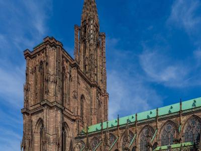 The image shows the Cathédrale Notre-Dame de Strasbourg, a massive Gothic cathedral with distinctive reddish-pink sandstone walls, a single towering spire, and green copper roofing, set against a blue sky with tourists in the foreground plaza.