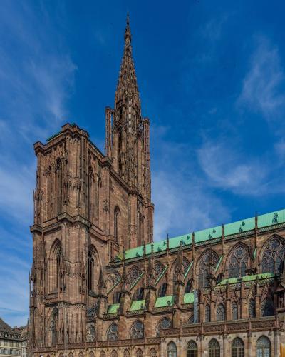 The image shows the Cathédrale Notre-Dame de Strasbourg, a massive Gothic cathedral with distinctive reddish-pink sandstone walls, a single towering spire, and green copper roofing, set against a blue sky with tourists in the foreground plaza.