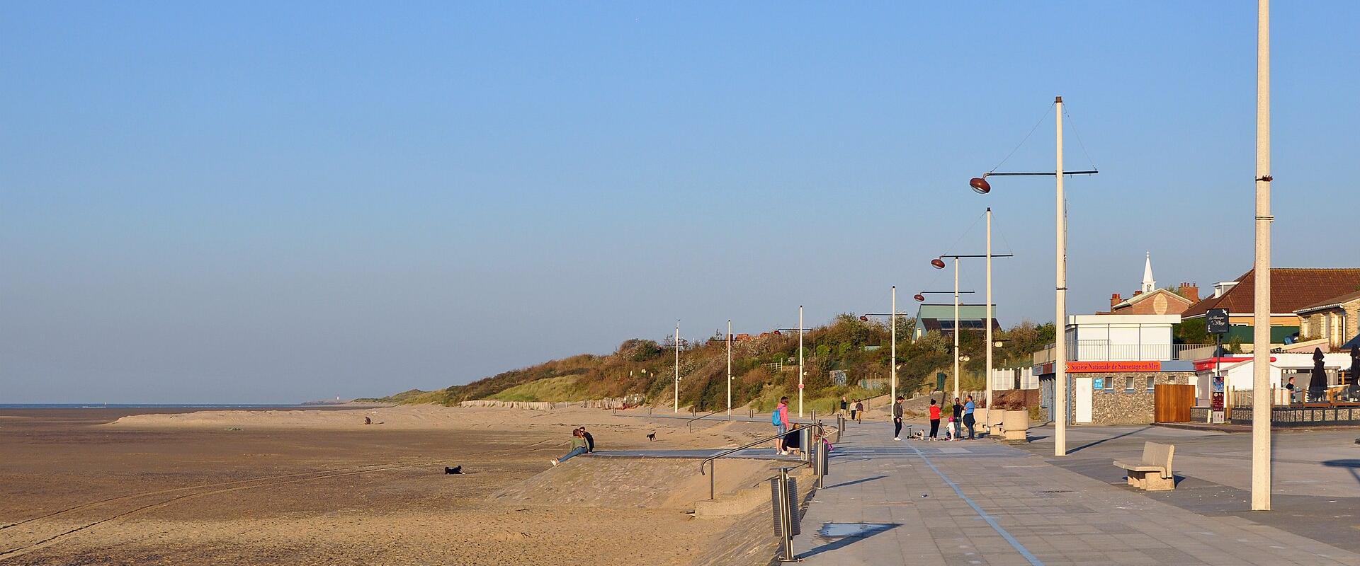 A wide sandy beach at low tide with a paved promenade, modern streetlights, benches, and a few people; buildings and a church steeple are visible in the background under a clear blue sky.