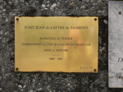 A close-up photograph of a brass commemorative plaque mounted on a pebbled concrete surface, inscribed with "PONT JEAN de LATTRE de TASSIGNY" and details about Marshal de Lattre de Tassigny.