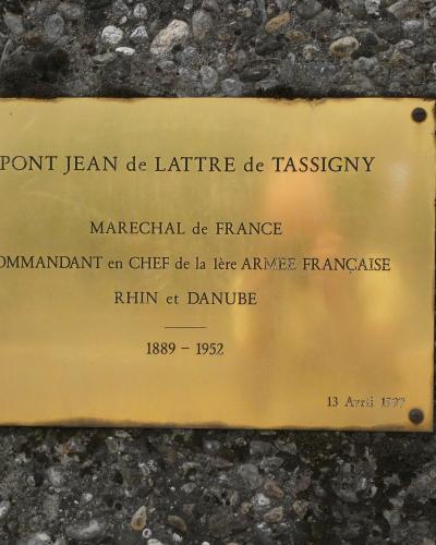 A close-up photograph of a brass commemorative plaque mounted on a pebbled concrete surface, inscribed with "PONT JEAN de LATTRE de TASSIGNY" and details about Marshal de Lattre de Tassigny.