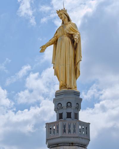 A golden statue of the Virgin Mary crowned and robed, standing atop a stone lantern and dome structure against a blue sky with white clouds.